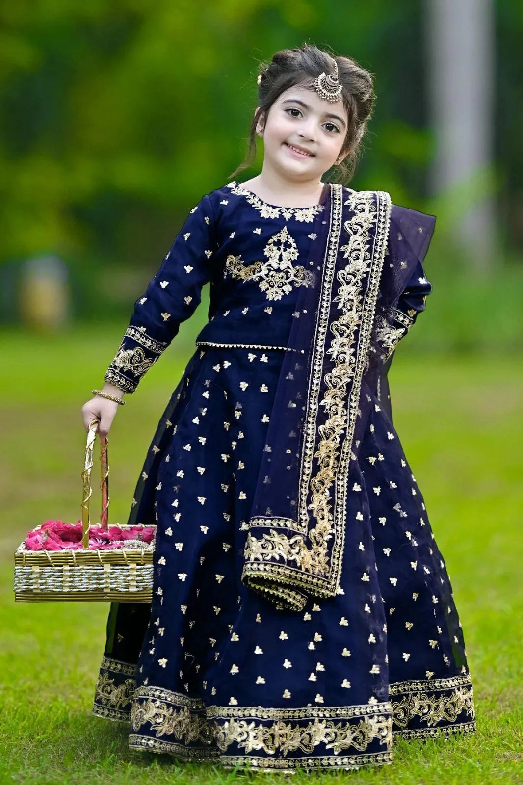 Young girl in a traditional navy blue outfit with gold embroidery, standing outdoors on grass.